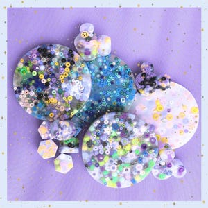 May include: A collection of colorful, handmade resin ornaments with embedded beads and glitter. The ornaments are round and hexagonal, featuring shades of blue, purple, green, and yellow. Some have small, decorative toppers.