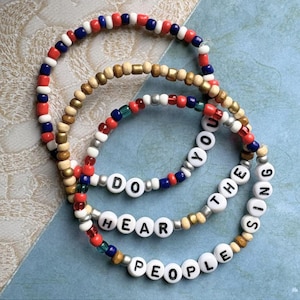 May include: Set of four beaded bracelets with the words "Do you hear the people sing" spelled out in white beads. The bracelets are made with red, white, blue, gold, and green beads.