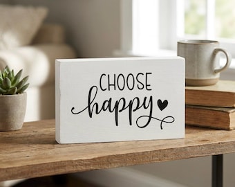 May include: A white wooden block with the words "CHOOSE happy" in black script and a heart symbol. The block is on a wooden table, with a mug and books in the background. A small succulent plant is also visible.