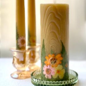 May include: Two tall yellow taper candles in clear glass holders and one thick yellow pillar candle with a wood grain texture, decorated with pressed orange, pink, and yellow flowers and green leaves, resting on a green glass plate.