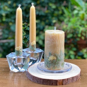 May include: Three candles with floral designs. Two tall candles are in crystal holders on a wooden surface. The third candle is a pillar candle with a wood grain design and is on a glass plate and a wooden base.