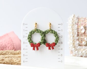 May include: A pair of festive earrings shaped like miniature wreaths. Each wreath is a ring of green, textured leaves, adorned with a small red bow. The earrings are displayed on a white stand with gold-colored posts.