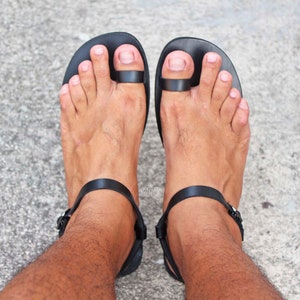 May include: Black leather sandals with a toe loop and ankle strap. The sandals are worn on a person's feet.