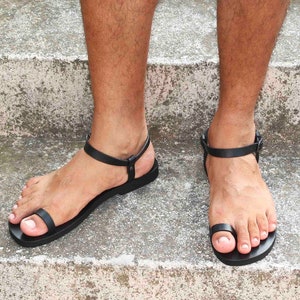 May include: A pair of black leather sandals with a thong style toe strap. The sandals have a simple design and are made of a soft, supple leather.