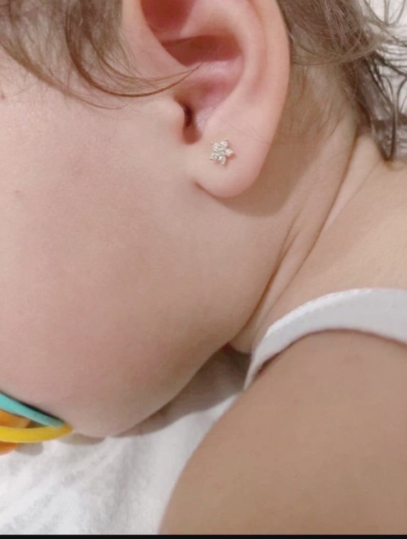 May include: A close-up of a baby's ear with a small, white flower-shaped earring.