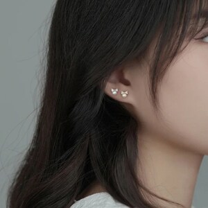 May include: A pair of silver earrings with a small pearl and a bow design. The earrings are shown on a model's ear.