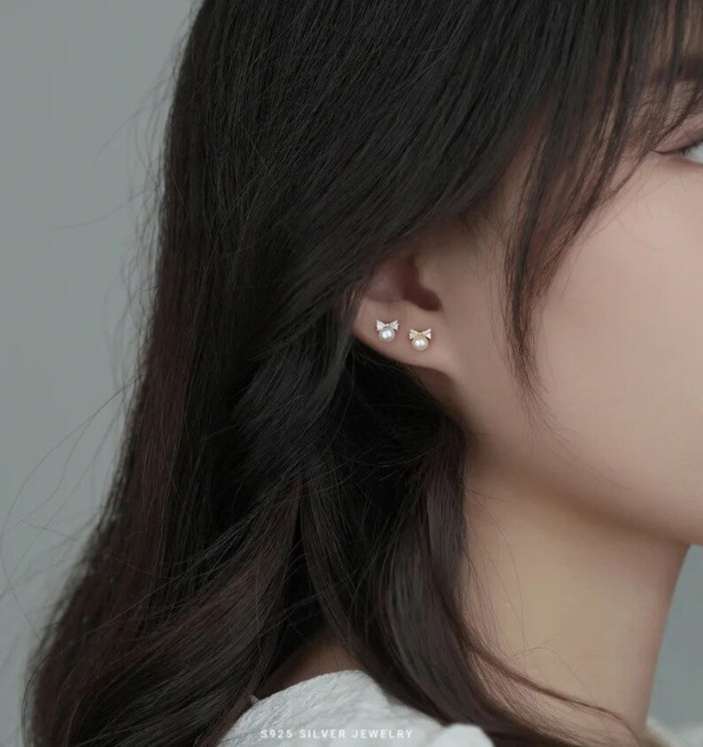 May include: A pair of silver earrings with a small pearl and a bow design. The earrings are shown on a model's ear.