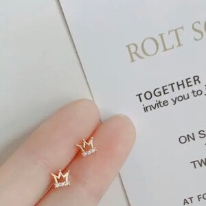 May include: A pair of gold crown earrings with small clear rhinestones. The earrings are shown on a person's ear.