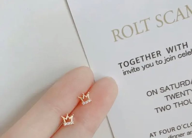 May include: A pair of gold crown earrings with small clear rhinestones. The earrings are shown on a person's ear.
