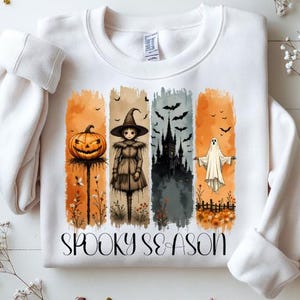 Puede incluir: Sudadera blanca con un gráfico colorido de Halloween. El diseño presenta una calabaza tallada, una bruja, un castillo embrujado y un fantasma, con las palabras "Spooky Season" en escritura negra. La obra de arte está sobre bloques de color verticales.