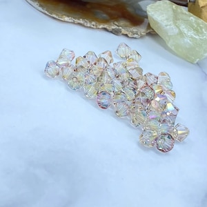 May include: A collection of iridescent crystal beads. The beads are faceted, reflecting light with a rainbow effect. They are scattered on a white surface, suggesting their use in jewelry making or crafts.