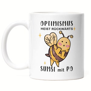 Può includere: Tazza in ceramica bianca con un'illustrazione di ape di cartone animato in nero e giallo. Il testo "Optimismus heisst rückwärts sumsi mit po" è stampato sulla tazza.