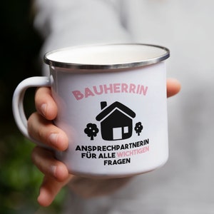 May include: White enamel mug with a pink and black design. The mug says "Bauherrin" and "Ansprechpartnerin f&uuml;r alle wichtigen Fragen".
