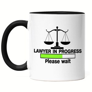 Könnte beinhalten: Eine weiße Kaffeetasse mit schwarzem Henkel und schwarzem Rand. Die Tasse zeigt eine schwarz-weiße Illustration einer Justiwaage und den Text "LAWYER IN PROGRESS" mit einem grünen Fortschrittsbalken und dem Text "Bitte warten".