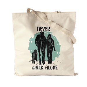 Könnte beinhalten: Eine weiße Canvas-Tragetasche mit einem türkisfarbenen Kreis und Silhouetten einer Person, eines Pferdes und eines Hundes, die gehen. Der Text "NEVER WALK ALONE" ist unter dem Kreis gedruckt.