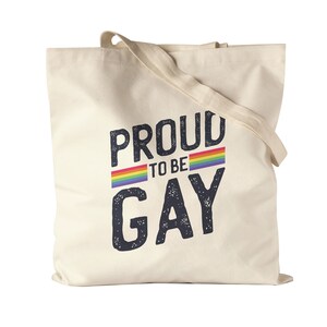 Op de afbeelding: Een beige canvas draagtas met de tekst "Proud to be Gay" in zwart met een regenboogstreep boven en onder de tekst.