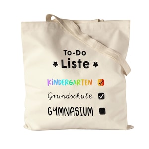 Può includere: Una borsa tote in tela bianca con il testo "To-Do Liste" e un elenco di controllo con le voci "Kindergarten", "Grundschule" e "Gymnasium". Le voci dell'elenco di controllo sono contrassegnate da segni di spunta e da un quadrato.