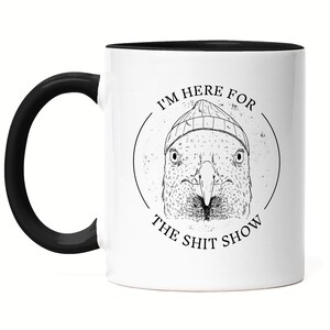 Peut inclure: Une tasse à café en céramique blanche avec une anse et un bord noirs. La tasse présente une illustration en noir et blanc d'un oiseau portant un bonnet. Le texte "I'm here for the shit show" est imprimé en cercle autour de l'oiseau.
