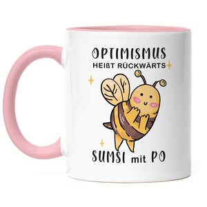 Può includere: Tazza in ceramica bianca con bordo e manico rosa. La tazza presenta un'ape di cartone animato con il testo "Optimismus heißt rückwärts sumsi mit po" in nero.