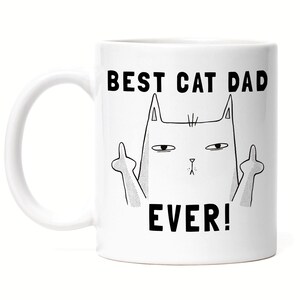 Może przedstawiać: Biały ceramiczny kubek do kawy z czarnym konturem kota pokazującego środkowy palec z nadrukowanym tekstem "BEST CAT DAD EVER!"