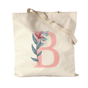 Op de afbeelding: Een natuurlijke canvas tote bag met een roze letter "B" en een aquarel bloemenontwerp. Het bloemenontwerp bevat blauwe bladeren en een roze bloem.
