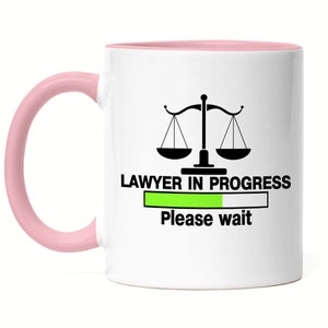 Könnte beinhalten: Eine weiße Kaffeetasse mit rosafarbenem Rand und Henkel. Die Tasse zeigt eine schwarz-weiße Grafik einer Justiwaage mit dem Text "LAWYER IN PROGRESS" und einem grünen Fortschrittsbalken mit dem Text "Bitte warten" darunter.