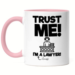 Op de afbeelding: Witte keramische koffiemok met een roze rand en handvat. De mok heeft zwarte tekst die "Trust Me! I'm a Lawyer! almost" luidt. Er is een zwart silhouet van een persoon die aan een bureau zit met boeken.