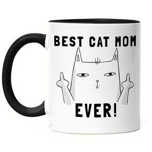 Może przedstawiać: Biały ceramiczny kubek do kawy z czarnym rantem i uchwytem. Kubek ma czarno-białą ilustrację kota pokazującego środkowy palec z nadrukowanym tekstem "BEST CAT MOM EVER!"