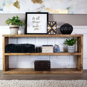 May include: A rustic wooden shelf unit with two shelves. The top shelf has a black framed print with the text "do all things with love", a black and gold geometric wooden wall decor, a black ceramic vase, and a potted plant. The bottom shelf has a black faux fur throw, a stack of books, a black box, and a potted plant.