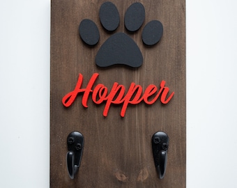 May include: A wooden dog leash holder with two hooks. The holder features a black paw print and the name "Hopper" in red letters.