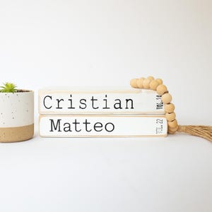 May include: Two white wooden blocks stacked on top of each other. The top block says "Cristian" and the bottom block says "Matteo". A wooden bead garland with a tassel is next to the blocks.