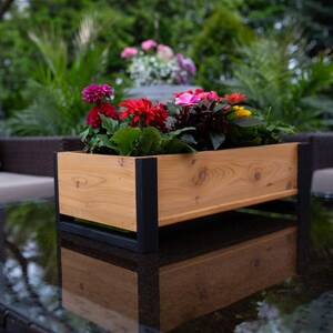 May include: A rectangular wooden planter box with a natural wood grain finish, supported by black metal legs. The planter is filled with vibrant red, pink, and yellow flowers and green foliage. The planter is sitting on a glass table.