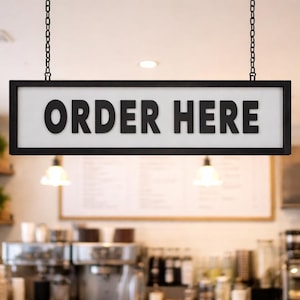 May include: A black-framed sign with the text "ORDER HERE" in bold, black letters hangs from chains. The sign has a white background. The background is a blurred interior of a cafe with lights and equipment.