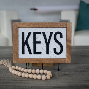 May include: A wooden sign with the word "KEYS" in black lettering on a white background. The sign is on a grey wooden table with a wooden bead garland in front of it.