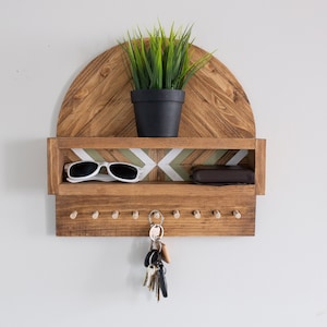 May include: A wooden key holder with a shelf and a geometric pattern. The key holder has a small plant on top and a pair of sunglasses and a wallet on the shelf. There are hooks on the bottom for hanging keys.
