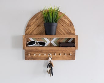 May include: A wooden key holder with a shelf and a geometric pattern. The key holder has a small plant on top and a pair of sunglasses and a wallet on the shelf. There are hooks on the bottom for hanging keys.