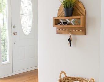 May include: A wooden wall-mounted shelf with a key rack and a small shelf for sunglasses. The shelf has a decorative chevron pattern and holds a potted plant. Keys hang from the key rack. The shelf is mounted on a white wall.