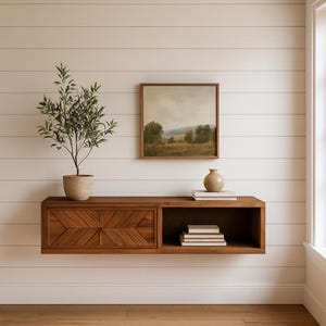 May include: A modern, floating wooden shelf with a geometric design on the left door. The shelf holds a small potted plant, a framed landscape painting, a vase, and books. The wall is white with horizontal wood paneling.