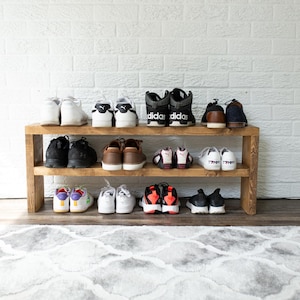 Peut inclure: Un porte-chaussures en bois à deux étagères, contenant une variété de baskets et de chaussures. Le porte-chaussures est contre un mur en briques blanches.