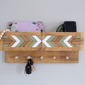 May include: Wooden key holder with a shelf and five hooks. The shelf has a geometric pattern of green, brown, and white painted arrows. There are keys, a phone, and sunglasses on the shelf.