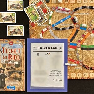 May include: A Ticket to Ride: Amsterdam board game set up for play. The game board is a map of Amsterdam with train tracks and train carriages. There are also destination tickets, train carriages, and a score pad. The game is for 2-4 players.