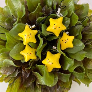 May include: Five yellow star-shaped charms with smiling faces and black cords. The charms are set against a backdrop of green and brown leaves. Each star has a silver-coloured clasp.