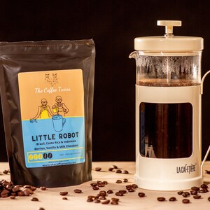 May include: A brown bag of coffee beans labelled "The Coffee Twins Little Robot" with a cartoon illustration of two people. The bag is sitting on a wooden surface with coffee beans scattered around it. A white French press with a glass carafe is filled with coffee and sitting on the wooden surface.