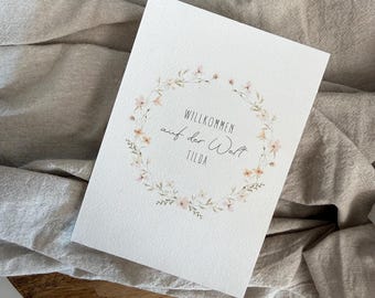 Könnte beinhalten: Eine weiße Karte mit dem Text "Willkommen auf der Welt Tilda", umgeben von einem floralen Kranz. Die Karte liegt auf einem Holztablett und einem beigen Leinentuch.