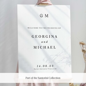 May include: A white wedding sign with black text announcing the wedding of Georgina and Michael. The sign includes the date "14.08.23" and the location "Saint Antonio Vineyard". The sign also includes the text "Part of the Santorini Collection".