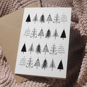 Könnte beinhalten: Eine weiße Grußkarte mit Illustrationen von schwarzen Weihnachtsbäumen, die jeweils mit einem goldenen Stern verziert sind. Die Karte wird von einem braunen Umschlag begleitet, vor einem strukturierten, hellrosa Hintergrund.