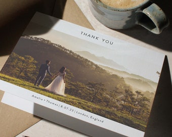 May include: A white thank you card with a photo of a couple in a field with mountains in the background. The text "THANK YOU" is printed in black at the top of the card. The text "Amelia + Thomas | 8.07.23 | London, England" is printed in black at the bottom of the card.