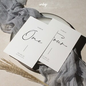Puede incluir: Dos tarjetas de mesa blancas con la escritura negra "One" y "Four". Cada tarjeta tiene una línea vertical y la fecha "05.08.21" con nombres. Las tarjetas están sobre un plato con tela gris y hierba seca.