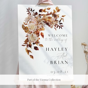 May include: A wedding welcome sign with a watercolor floral design in shades of brown, orange, and cream. The sign reads "Welcome to the wedding of Hayley and Brian 05.08.21 St. Tristans Church, Vienna. Part of the Vienna Collection."