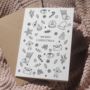 Könnte beinhalten: Eine weiße Weihnachtskarte mit schwarzen Strichzeichnungen von Feiertagsikonen, darunter ein Lebkuchenmann, ein Strumpf und eine Zuckerstange. Die Worte "MERRY CHRISTMAS" sind in der Mitte aufgedruckt. Die Karte wird von einem braunen Umschlag begleitet.
